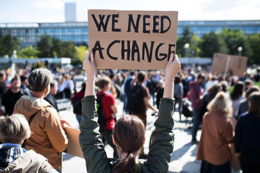 Valokuva mielenosoituksesta, jossa ihminen pitää käsissään kylttiä, jossa lukee teksti "We need a change".