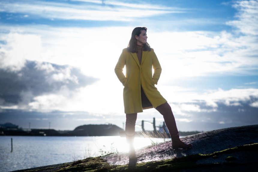 Marianne Heikkilä seisoo rantakalliolla keltaisessa takissa, ja aurinko paistaa hänen takaa.